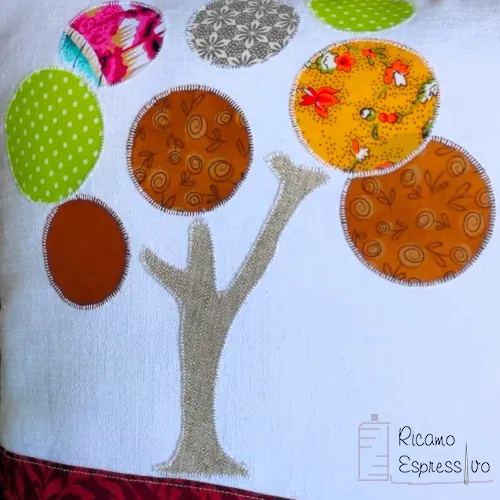 Cuscino artigianale in cotone e lino su cui si ha un albero in applique che sorge su un terreno stratificato da vari tessuti e da un fiore ricamato a mano.